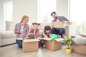 Young happy smiling family four persons unwrapping carton boxes with stuff in light living room, moving to new home, renovation