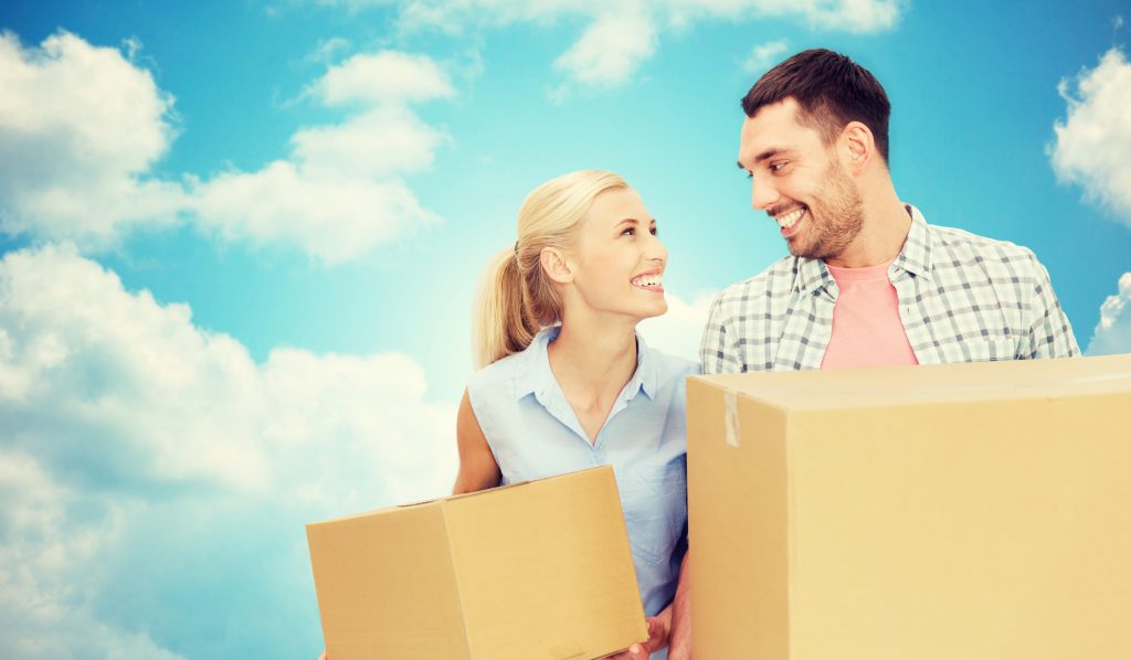 Blond woman and dark haired man standing close to each other, gazing at one another, each holding moving boxes with a bright blue sky and white clouds in the background