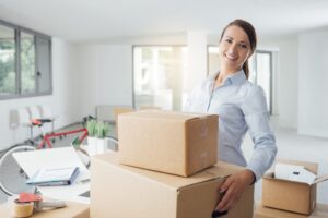 A young brown haired woman with hair in a pony tail standing in home office holding moving boxes