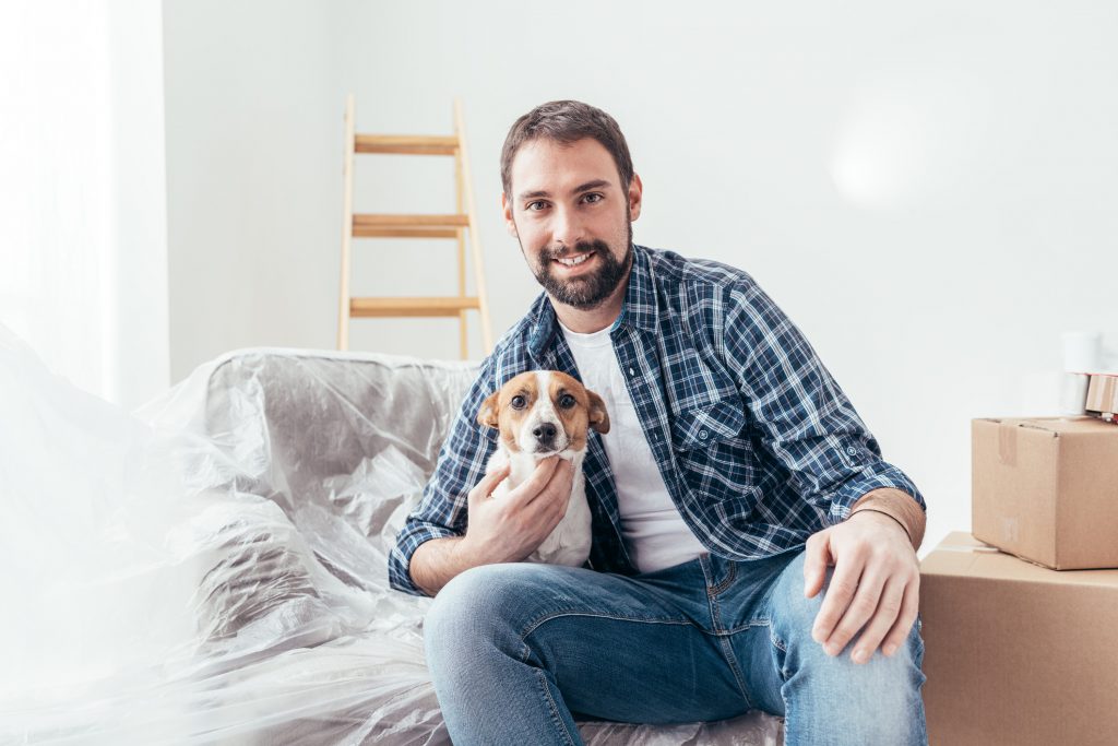 A man sitting on couch holding a small dog with moving boxes nearby and a ladder in the background.