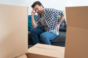 man in checkered shirt sitting on a couch holding his back in pain while surrounded by moving boxes
