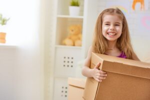 A young girl with long blonde hair smiling holding a moving box with a teddy bear on a shelf in the background