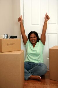 A woman in a green short sleeved short with her arms reached joyously in the air sitting on the floor next to moving boxes.