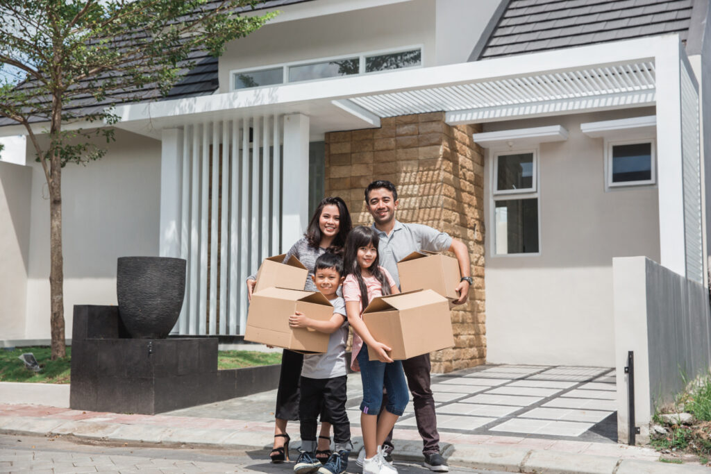 A family with man, woman, young, boy, young girl standing in front of house holding moving boxes.