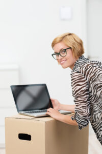 Young woman using a laptop on a cardboard box as she catches up on news in her new home turning to smile at the camera