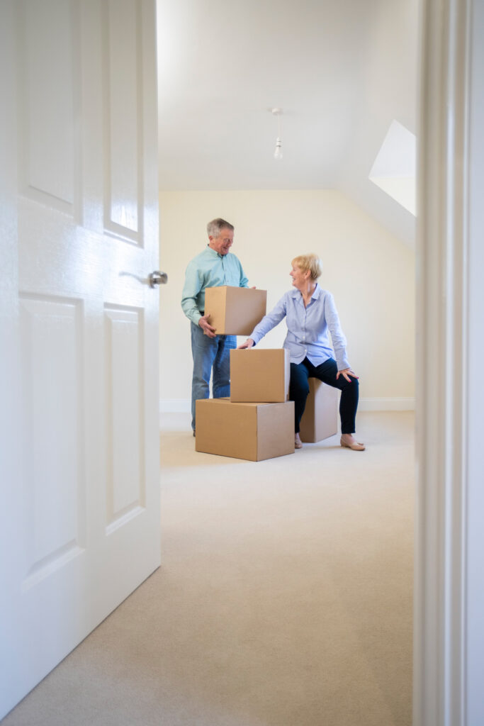 Senior couple downsizing in retirement carrying boxes into new home on moving day.