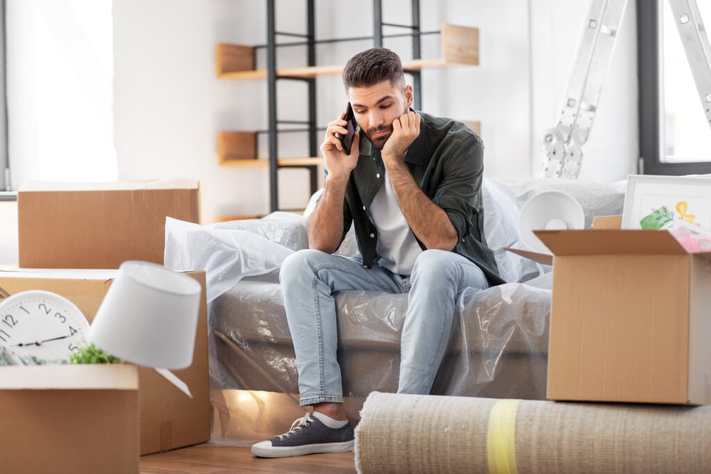 Moving, people and real estate concept - man thinking and listening with boxes calling on smartphone at new home