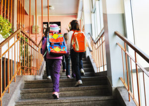 Children with backpacks on at a school walking up stairs.