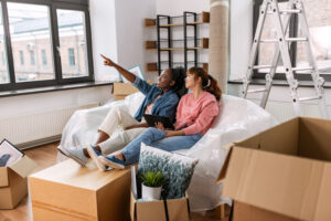 Two women sitting on a sofa surrounded by moving boxes. one woman is pointing out the window as the other woman looks.