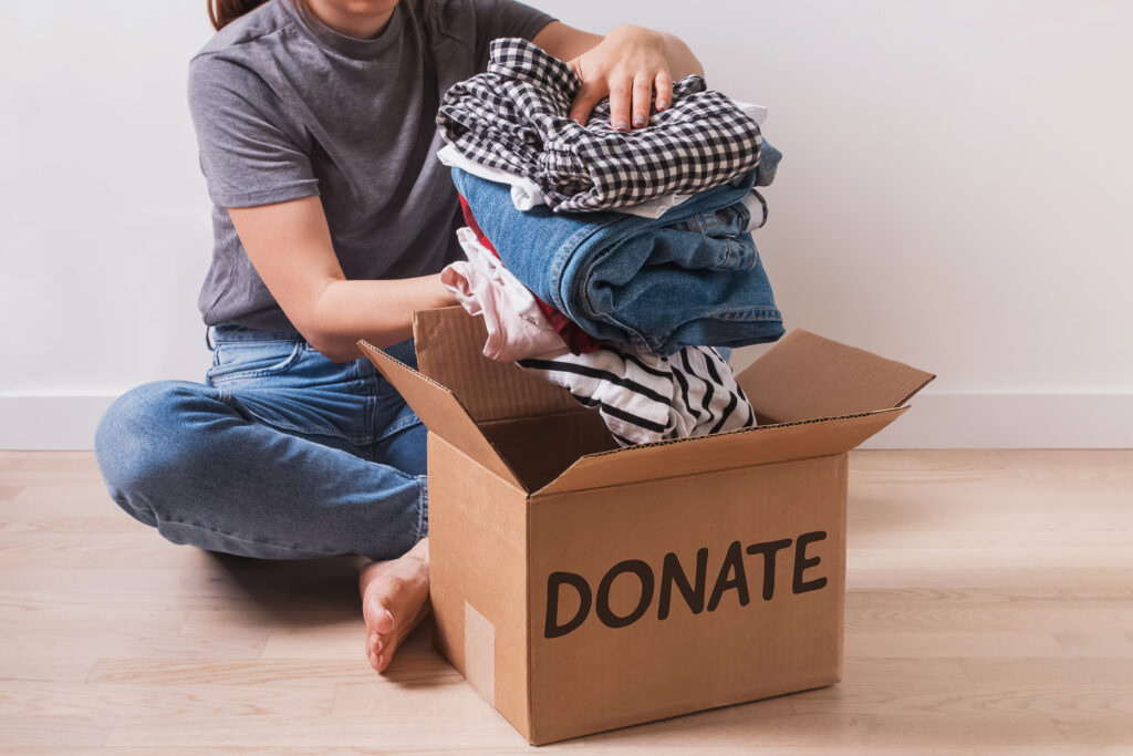 Person sitting on floor placing folded clothes into a cardboard box labeled donate.
