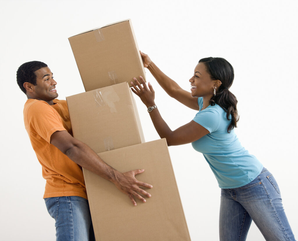 Man in an orange shirt holding a stack of moving boxes with woman in a blue shirt trying to help him balance the boxes.