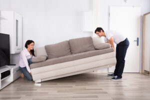 Woman at one end of a large couch trying to lift it and a man at the other end also lifting it.