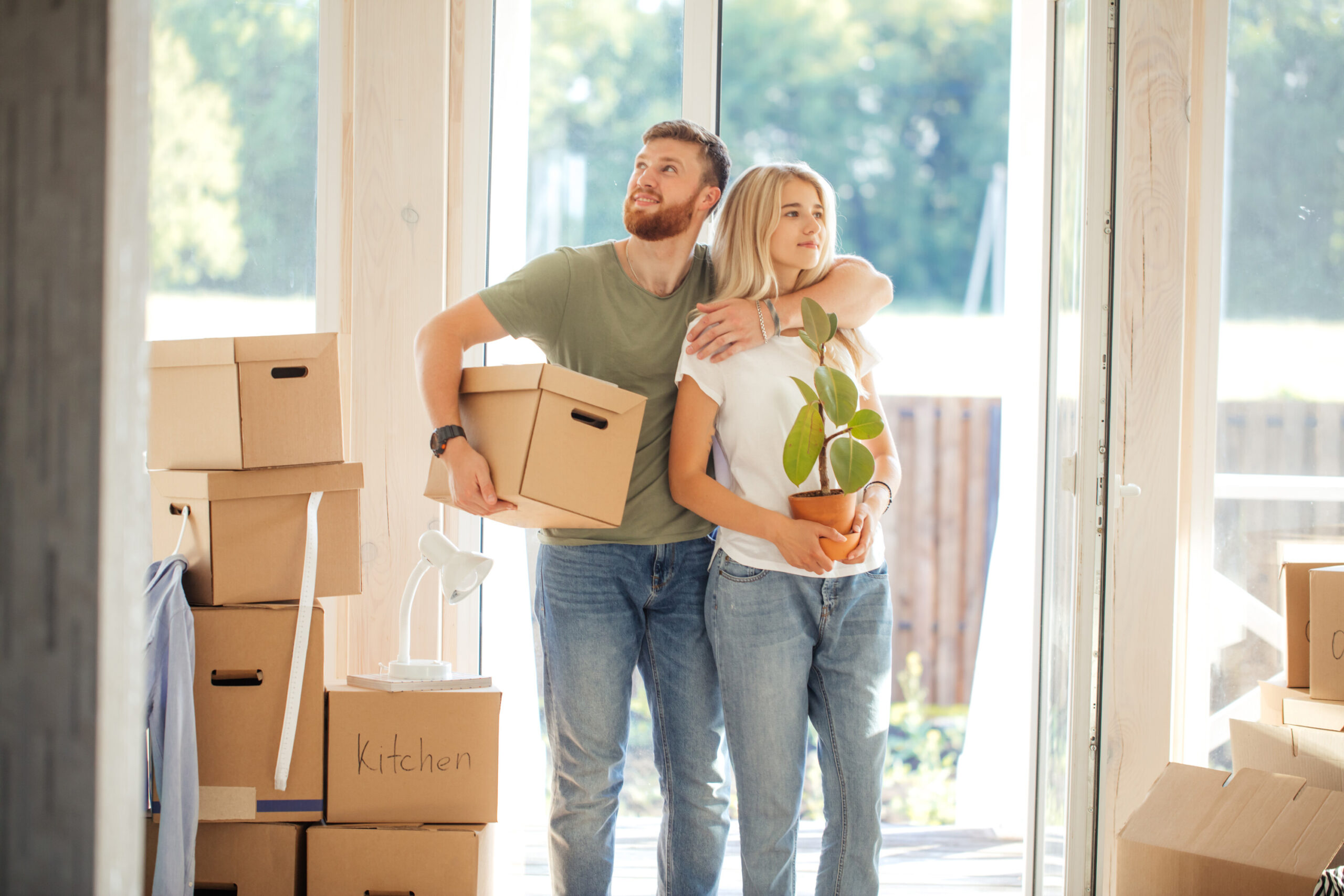 A man holding a moving box and a woman holding a plant embrace, standing in front of moving boxes and look around their home.