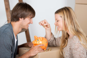Woman and man putting money in a piggy bank with moving boxes in the background.