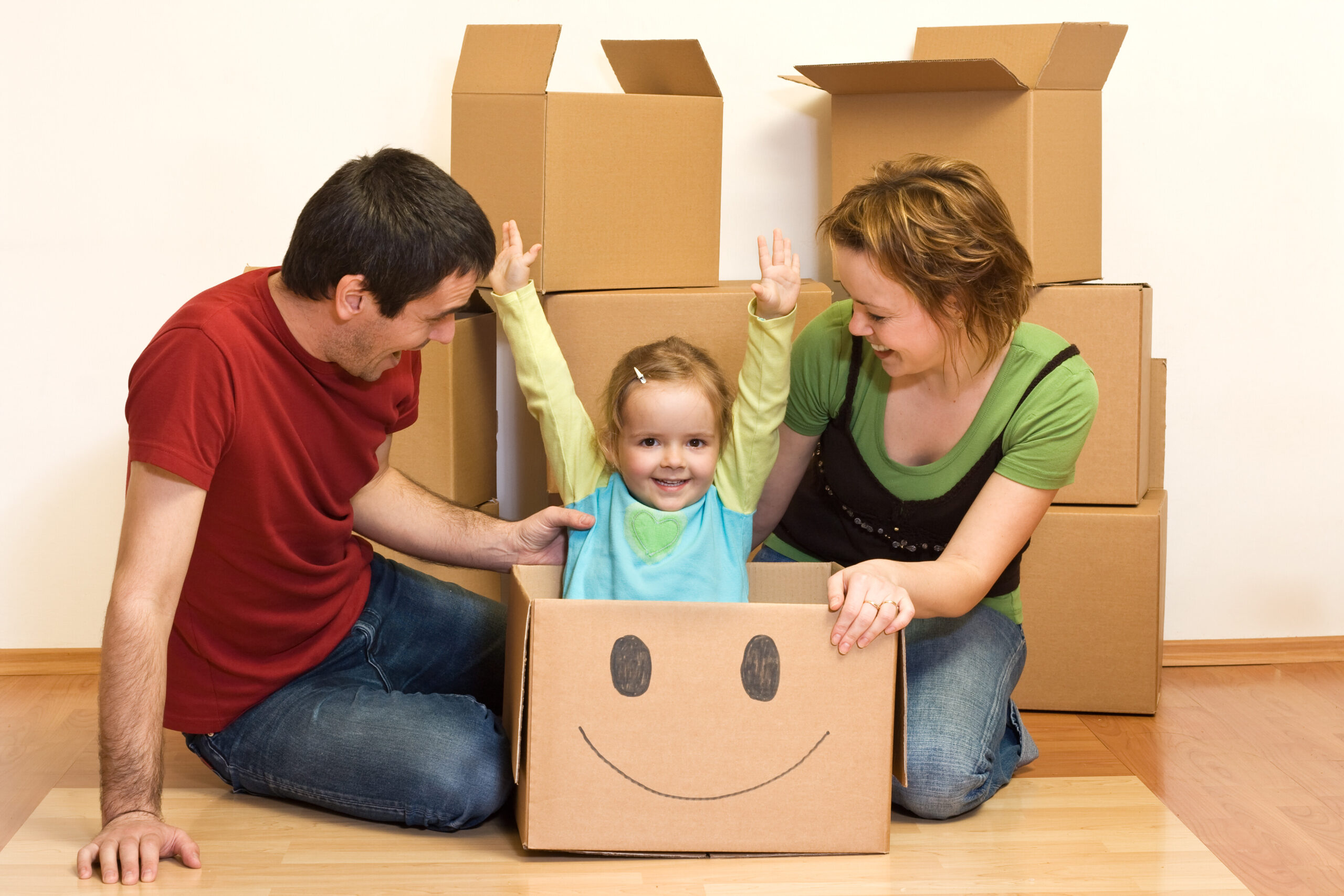 Small child in a moving box with a smiley face on the side. Attentive parents are next to her.