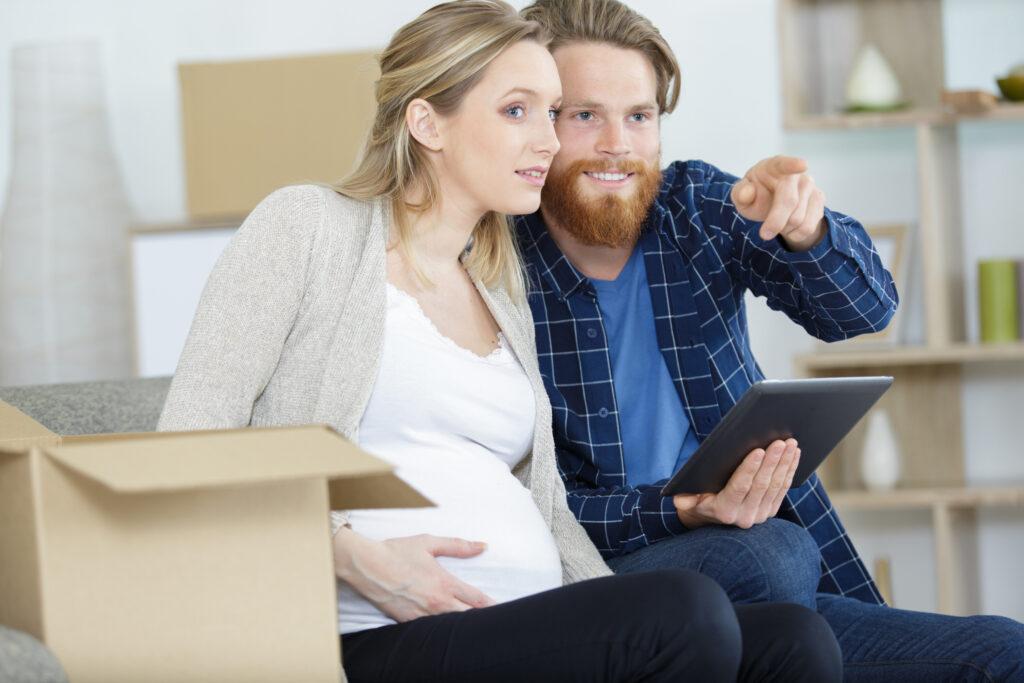 Expectant woman and man sit on a couch in front of moving boxes preparing for a move.