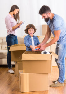 Young boy packing a moving box with the help of his dad with mom and other moving m boxes in the background